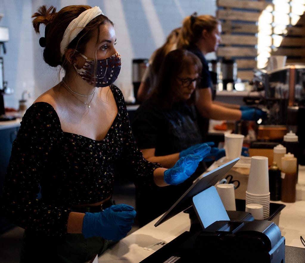 One masked woman behind a coffee counter beside two unmasked women.