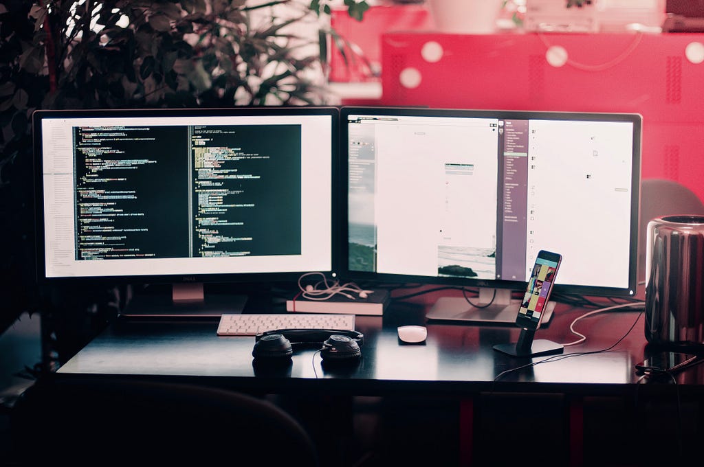 A desk holding two monitors with code on the screens, a smartphone, headphones, and a keyboard