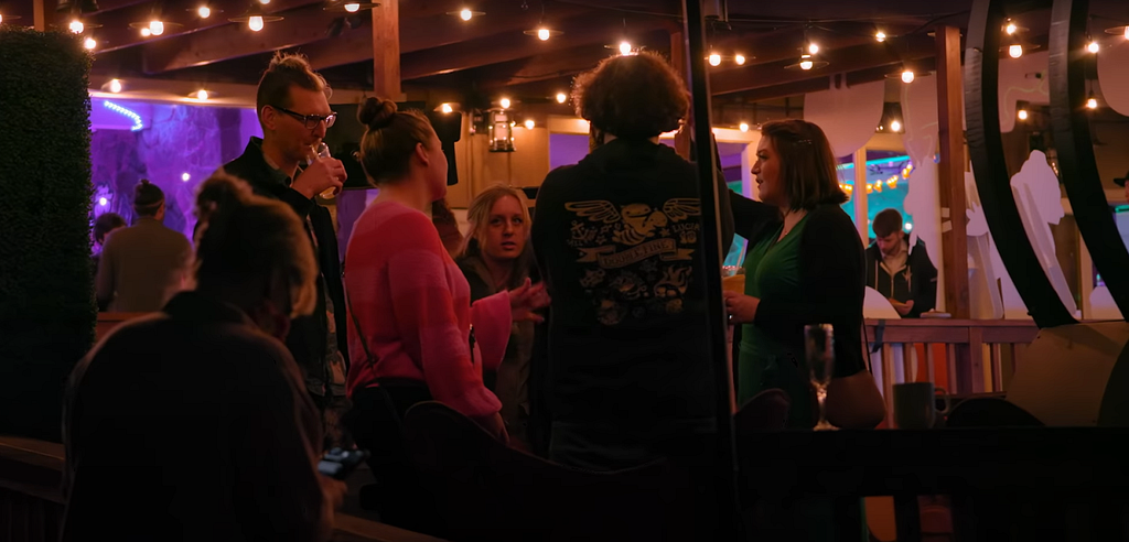 Double Fine employees gather in a circle talking to one another, with their backs to the camera. They are on a charmingly lit back porch at night during their release party, with warm string lights above their heads