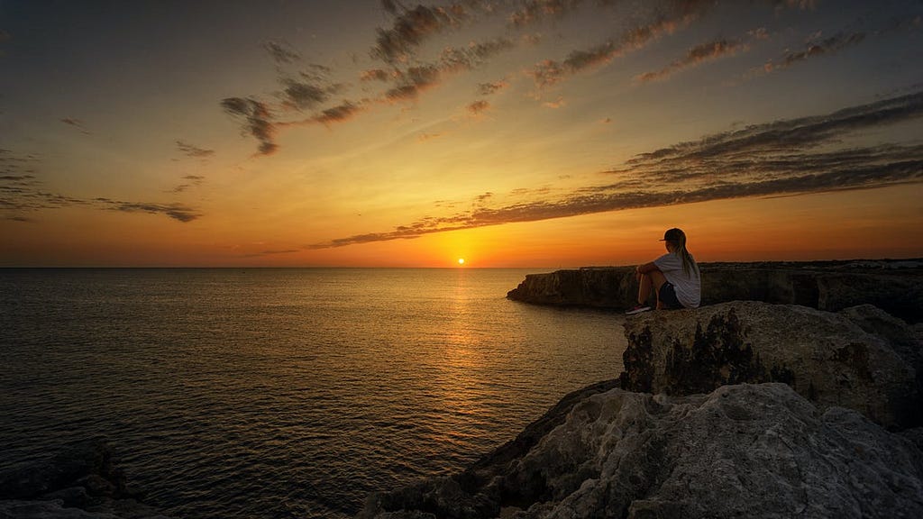 person sitting on a cliff looking at still water during a beautiful sunset