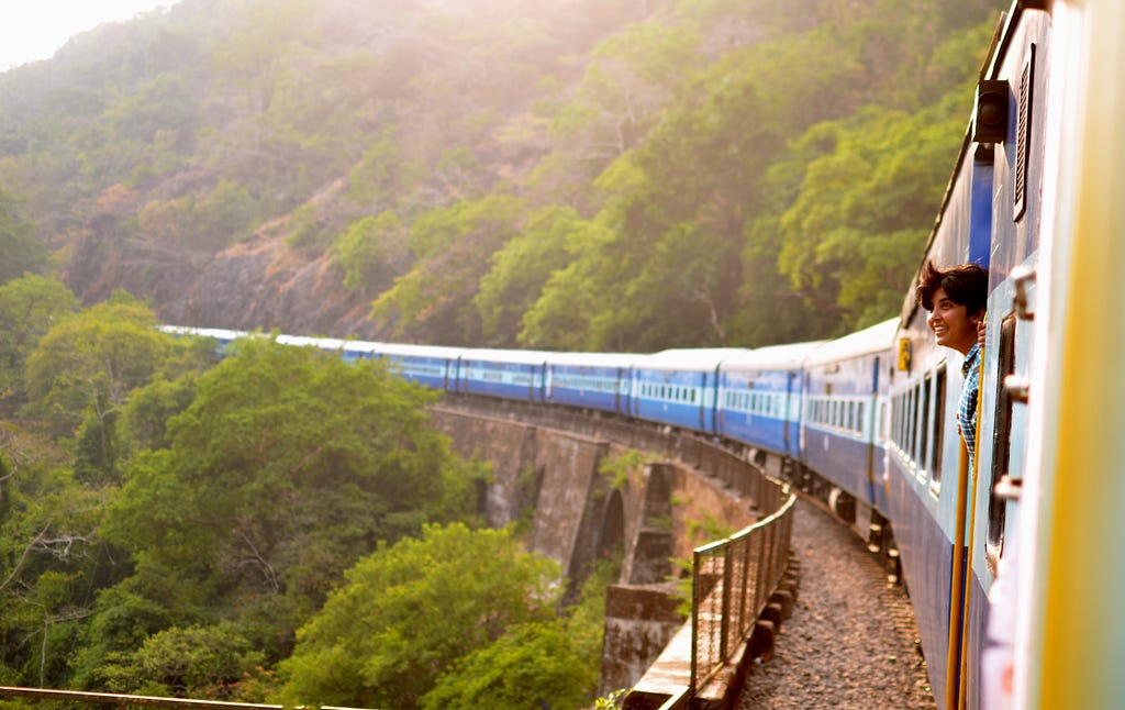 A person rides a train through the Indian countryside and sticks their head out of the window, smiling.