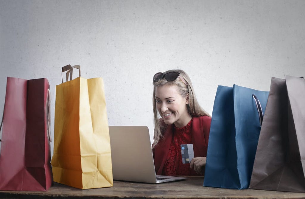 An ecstatic person is seen in front of a laptop holding a credit card. They are surrounded with multiple shopping bags.