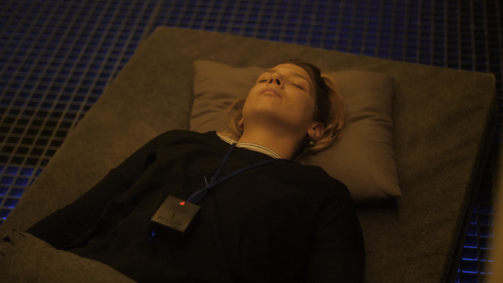 A light skinned person is laying face up at rest while participating in an immersive art environment with sound and haptic elements. The person is on a gray mat, eyes closed with calm expression, while a small electronic device component in the installation is on a neck strap and laying over the person’s heart.