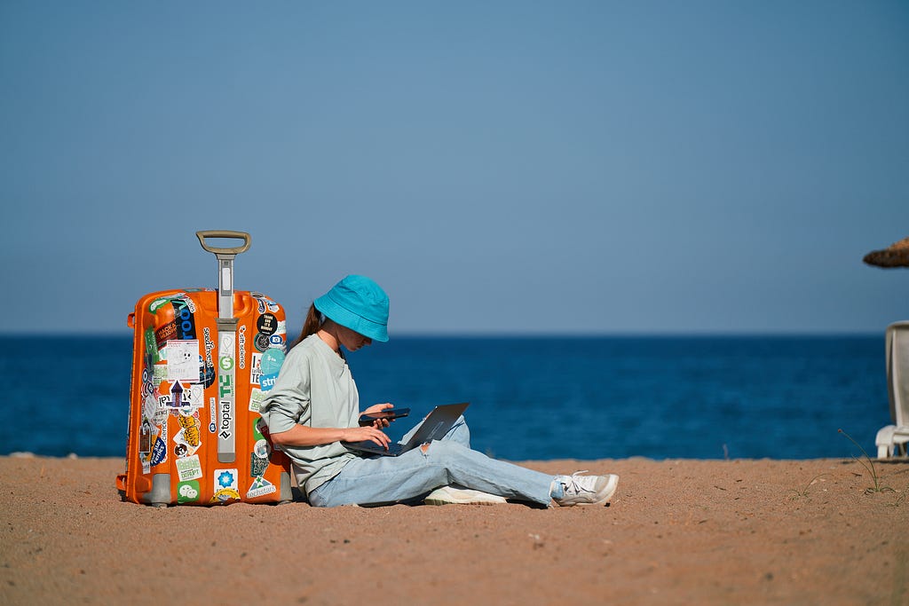 Uma pessoa sentada na areia da praia com o mar no fundo e céu azul. Ela está encostada em uma mala de viagem e com o laptop no colo, enquanto olha o celular