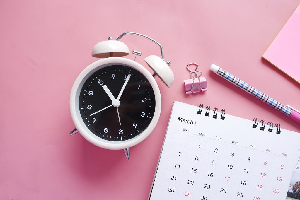 A white clock, calendar, pen, and pink clip on a pink background