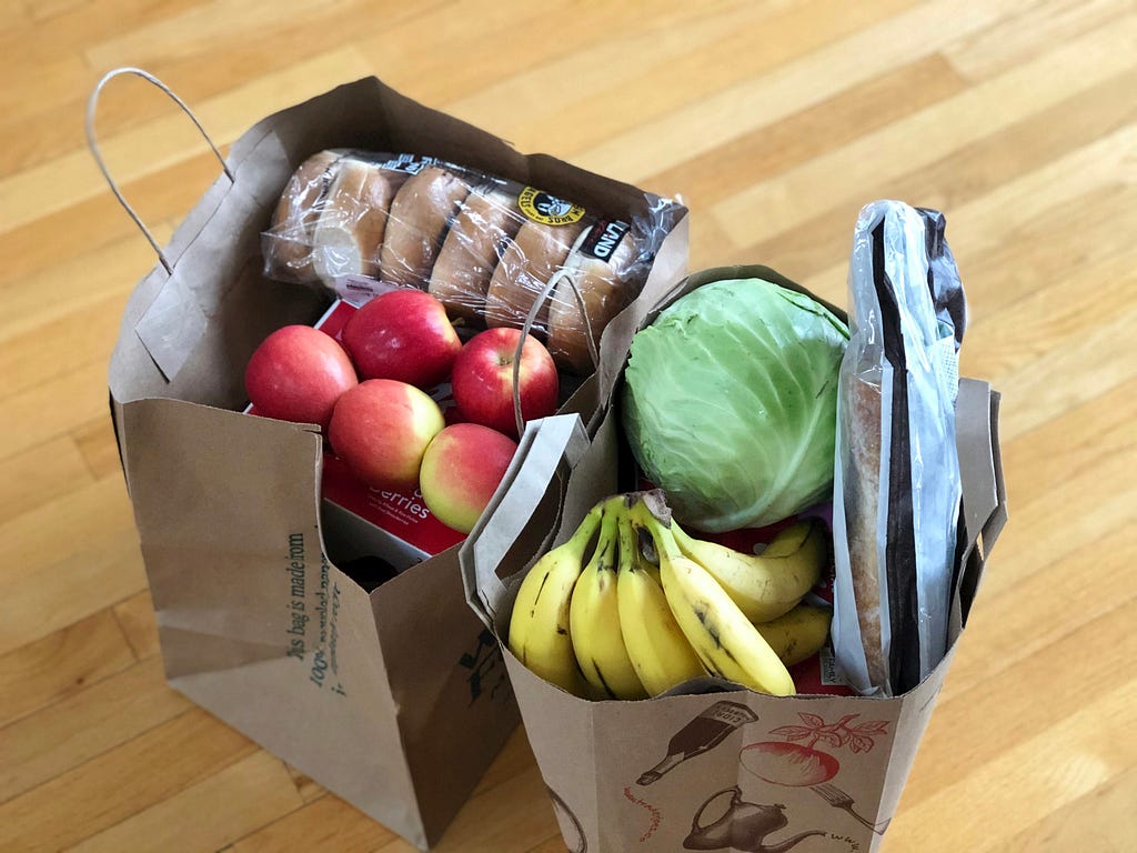 Two brown shopping bags filled with food such as bananas, red apples, bagels, iceberg lettuce and bread.