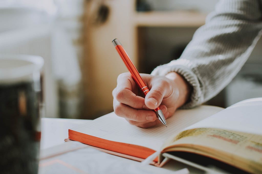 A person holds a bright orange pen over a notebook, their hand poised to write.