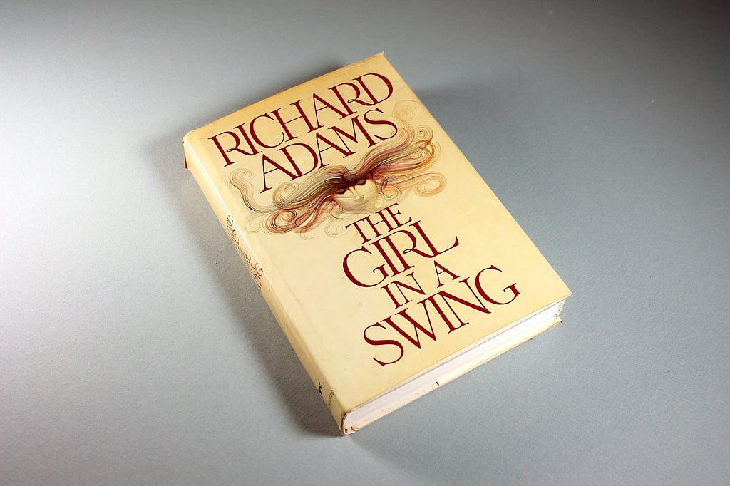 Another image of The Girl in a Swing by Richard Adams. It’s also a hardcover copy and a first-edition, but the dust jacket art is different. It’s of a woman’s face. Her eyes are closed, and her blonde hair swirls around her head like she’s in water.