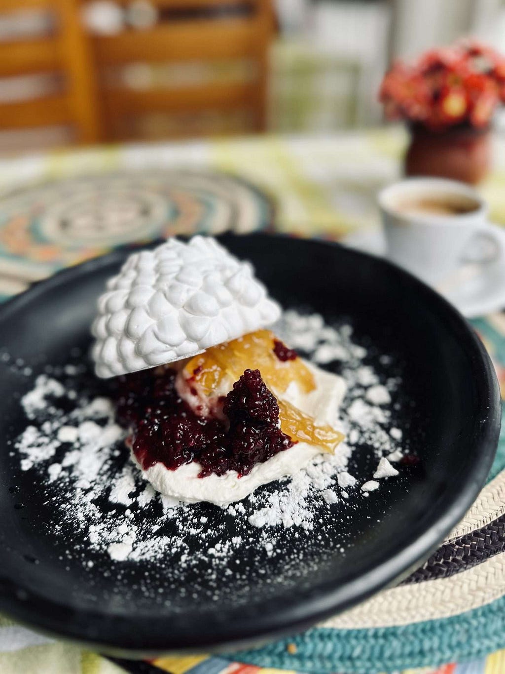Meruengón, a dessert with meringue, berry sauce, and fruit is dusted with powdered sugar on a black plate. A cup of coffee and a small flower arrangement are in the background.
