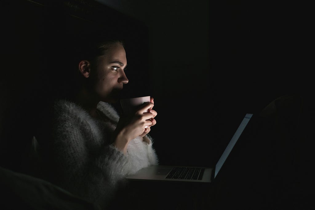 Woman looking at a laptop holding a drink in a dark room.