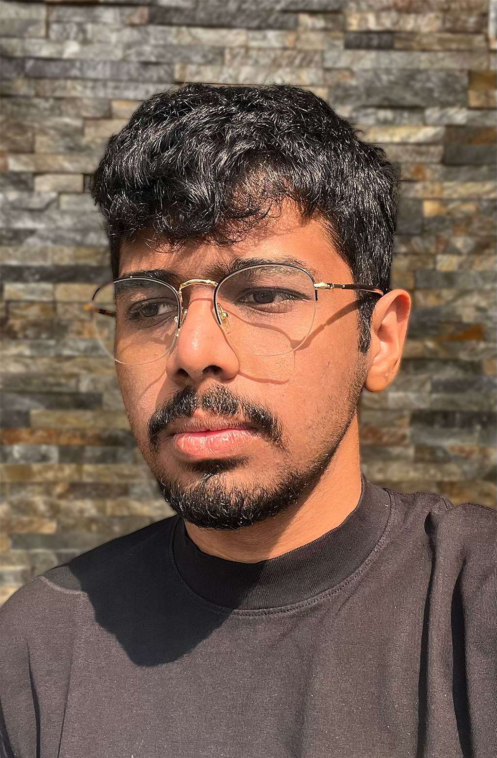 Photograph of an Indian man with black hair and glasses. He is standing in front of a stone wall, wearing a black t-shirt.