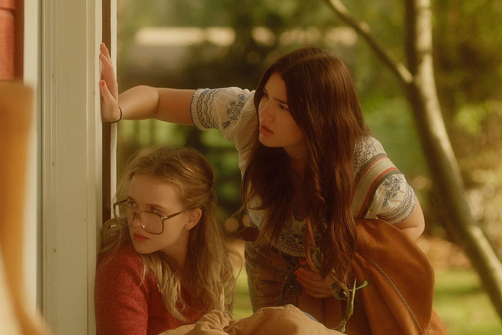 Kate and Tully, in their teenage versions, are shown spying through a window. Kate is sitting on the floor and Tully is bending over her with an arm on the wall, above Kate’s head. Her other arm is holding a brown coat. The scene has brown and green tones, thanks to Tully’s coat and Kate’s red-brownish jumper, and the green trees in the background.