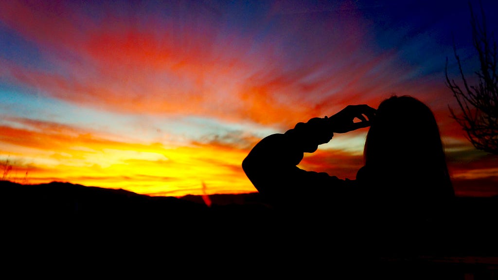 Black siloulette of woman looking at sunrise with her hand on here forehead.