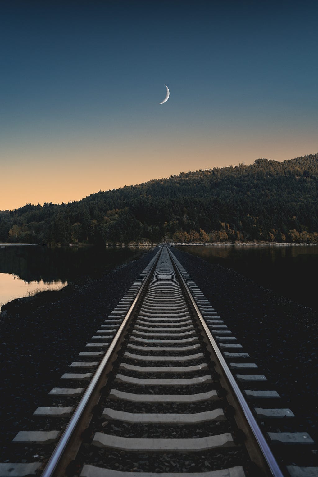 photo of train tracks leading to a forested hill or small mountain, under a golden-blue dusk sky with high crescent moon