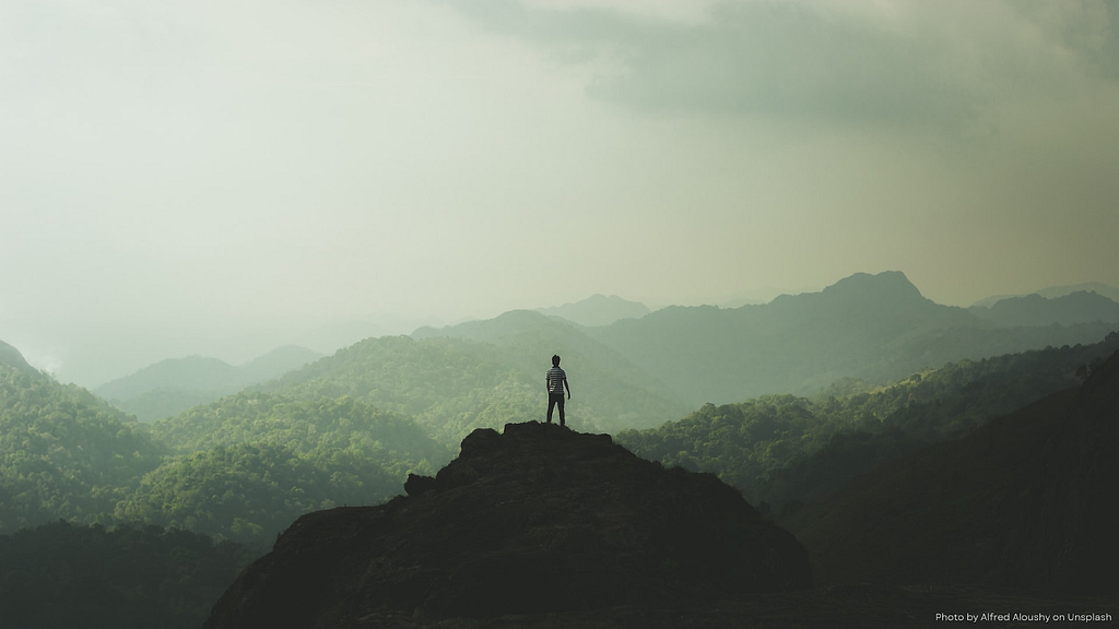 Photo of a lone male figure standing at the top of a rocky rise looking out over a series of mountain tops that gradually fade into the mist. Photo credit to Alfred Aloushy on Unsplash.