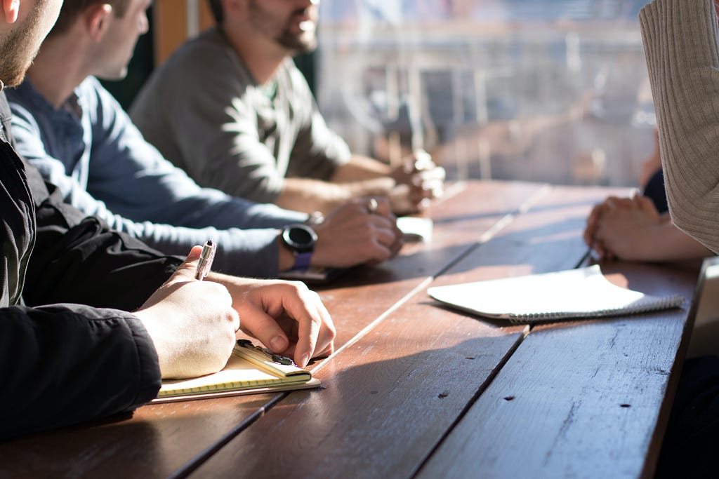 A group of people, with pens and paper, sitting at a table near a window