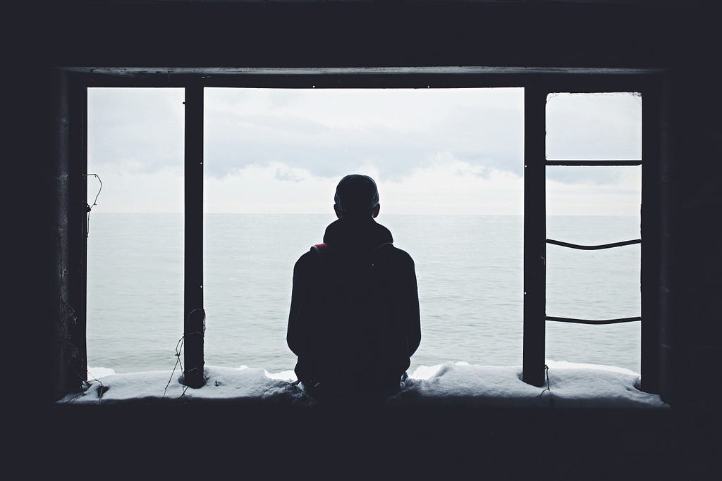 A guy sits alone at the edge of a window, watching the ocean view from a dark room.