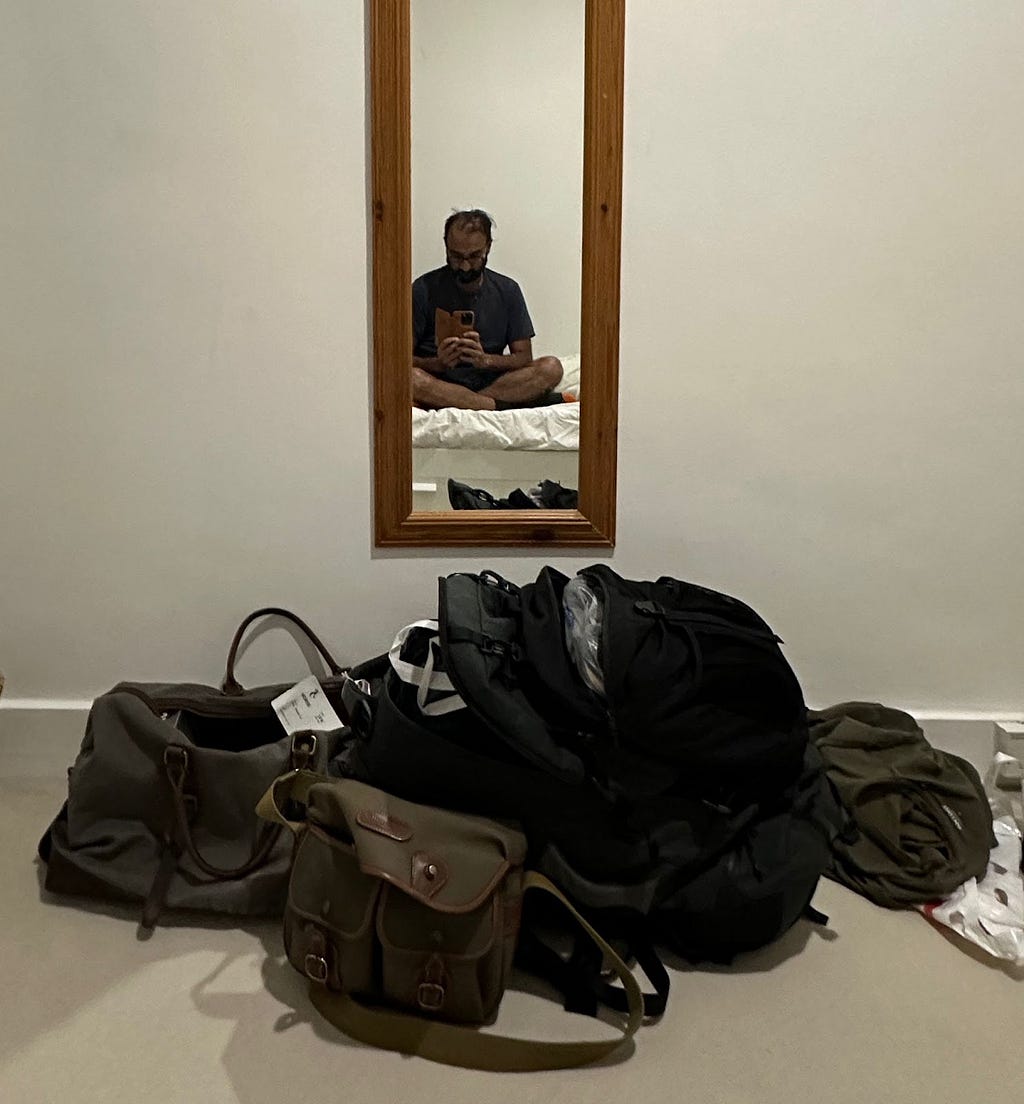 Bags open against wall with slim mirror reflecting a brown man with beard and glasses sat on bed cross-legged taking photo.