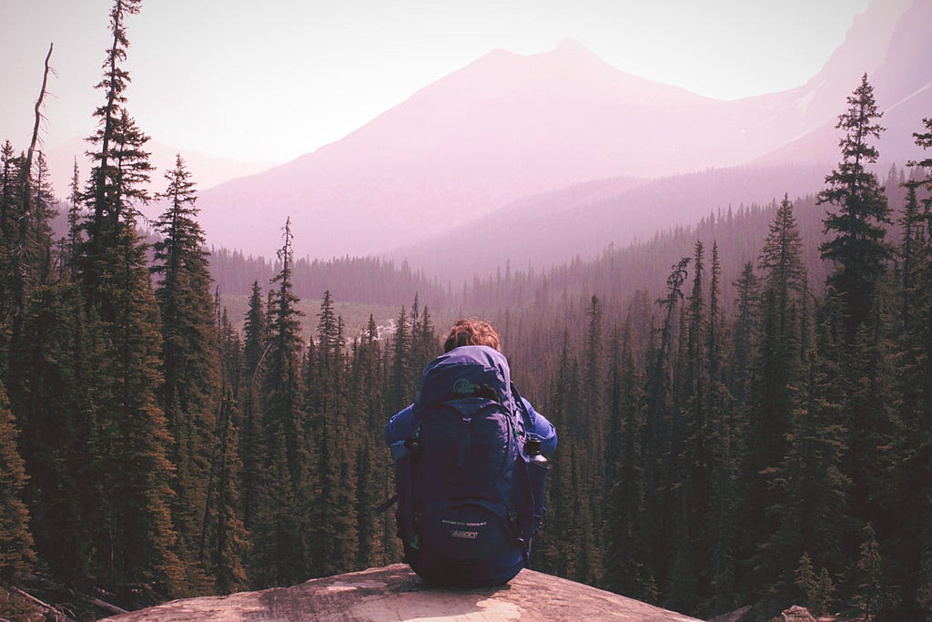 Uma fotografia de uma pessoa contemplando uma vista de floresta e montanha, assentado com sua mochila de trecking nas costas.