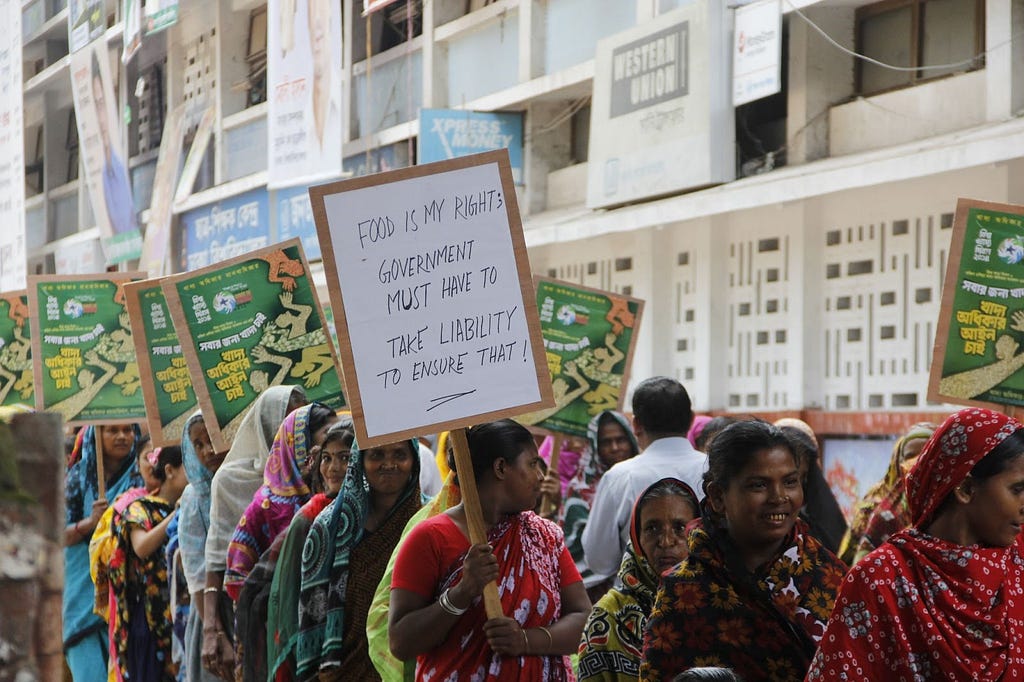 A line of women protesters pass by a building. One holds a sign that reads: “Food is my right; Government must have to take liability to ensure that!”