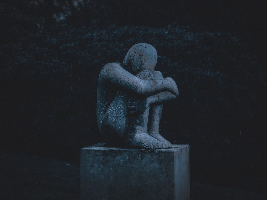A grey marble statue of a child resting its head on its arms, folded across its knees