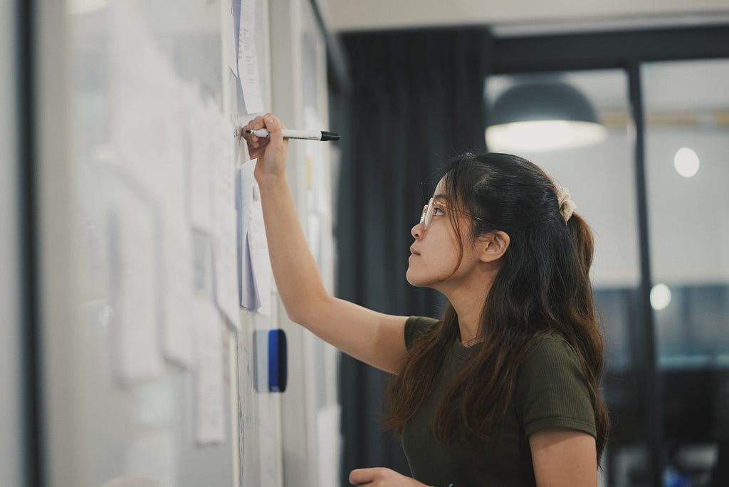 A young woman in a black t-shirt writes on a white board covered with sticky notes