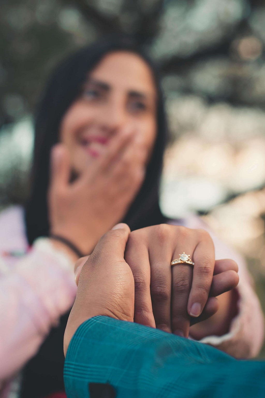 A close-up of a couple’s hands, with a focus on the woman’s hand showcasing an engagement ring. The background features the smiling, slightly blurred face of the woman, who has one hand covering her mouth in surprise or joy.