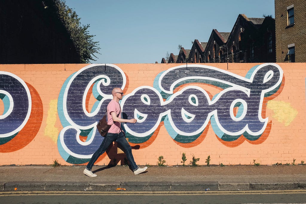 A man walks past a graffiti mural that says “Good” on a bright sunny day.