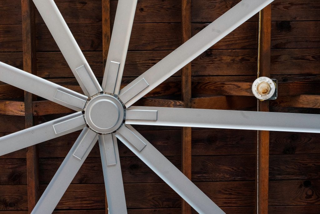 Nine bladed silver industrial fan against a wood ceiling.