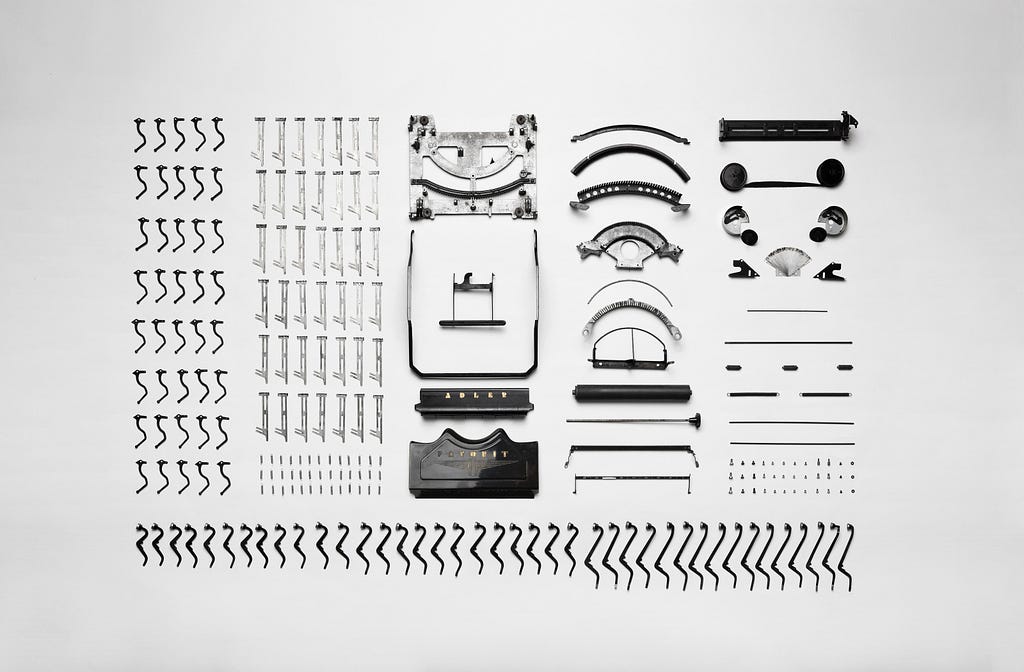 A black and white photo of a deconstructed typewriter, all the pieces laid out neatly, each similar type of part, e.g. letter keys, laid out in an ordered, tabular format. Neat, and aesthetically pleasing.