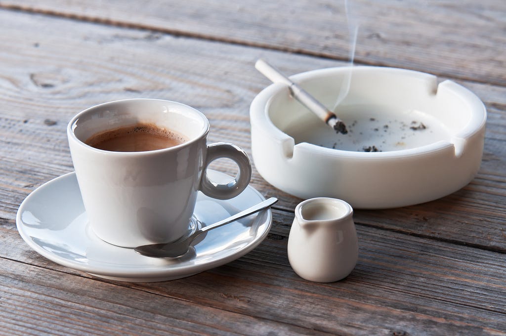 A cup of coffee and an ashtray with a smoldering cigarette on a wooden table