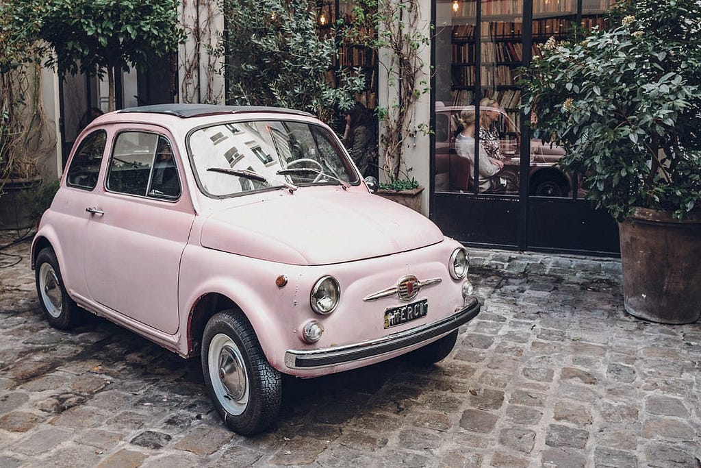 Imagen de un automóvil Fiat 500 clásico de color rosa pastel estacionado en un patio adoquinado. El vehículo está frente a una fachada de edificio con grandes ventanales y plantas, lo que sugiere un ambiente urbano y acogedor. La combinación del diseño retro del coche y el entorno tranquilo crea una escena encantadora y nostálgica.