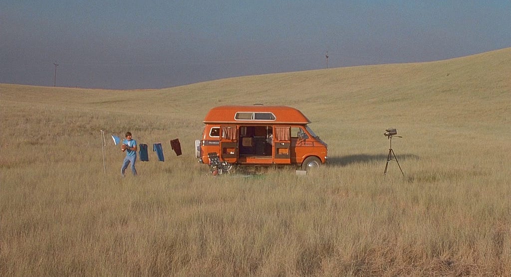 A man stands in a field next to the van he’s living in, holding a football, and recording himself with an old camcorder on a tripod.