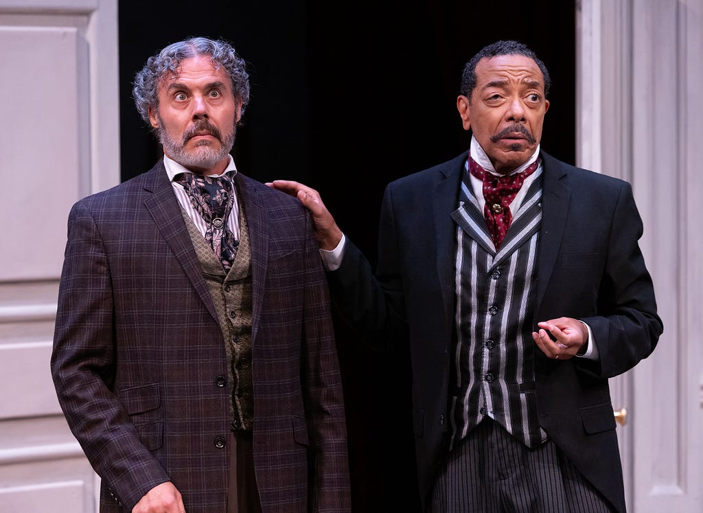 A man with curly gray hair and a beard wearing a brown suit and cravat looks wide-eyed while another man in a black jacket and striped vest lays a hand on his shoulder.