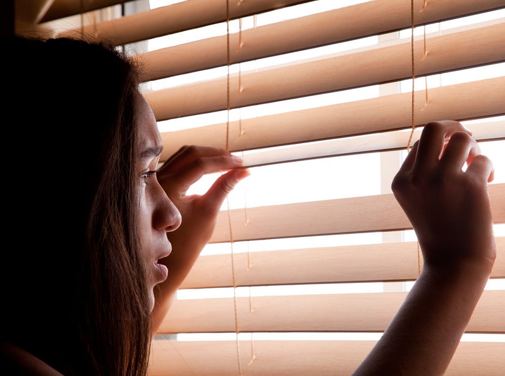 A woman looks out a window through blinds. Photo by lathuric/Getty Images