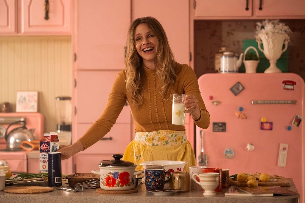 Cloud, Tully’s mom, is shown in a medium shot cooking in a fully-pink kitchen, with several ingredients on the counter. She is holding a glass of milk and there are several ingredients on the counter as if she was in the middle of cooking. She is wearing a mustard, long-sleeve top and a yellow apron with sunflowers on it.