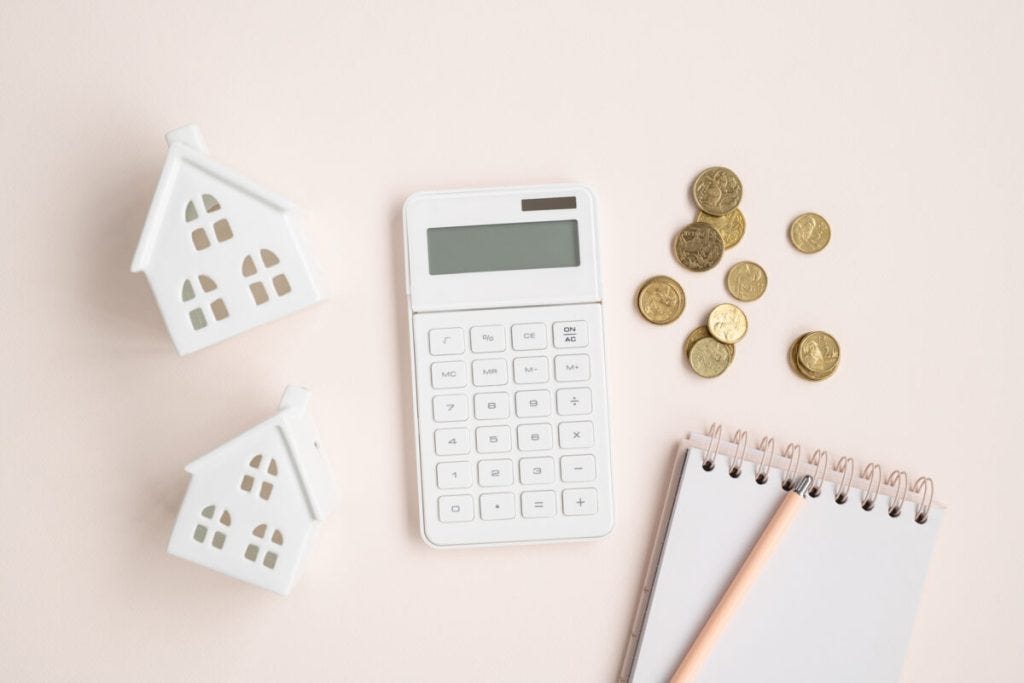 A flat lay of two small white ceramic house models, a white calculator, scattered coins, and a notebook with a pencil on a light pink background.