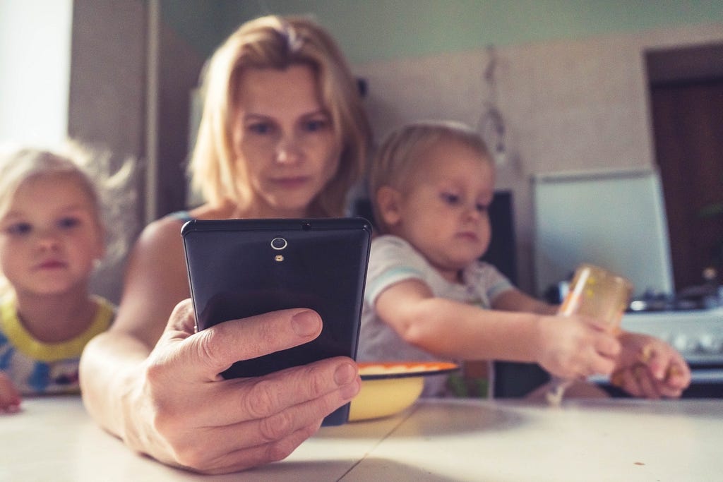 A mother sits at a table with her two young children, scrolling on her phone