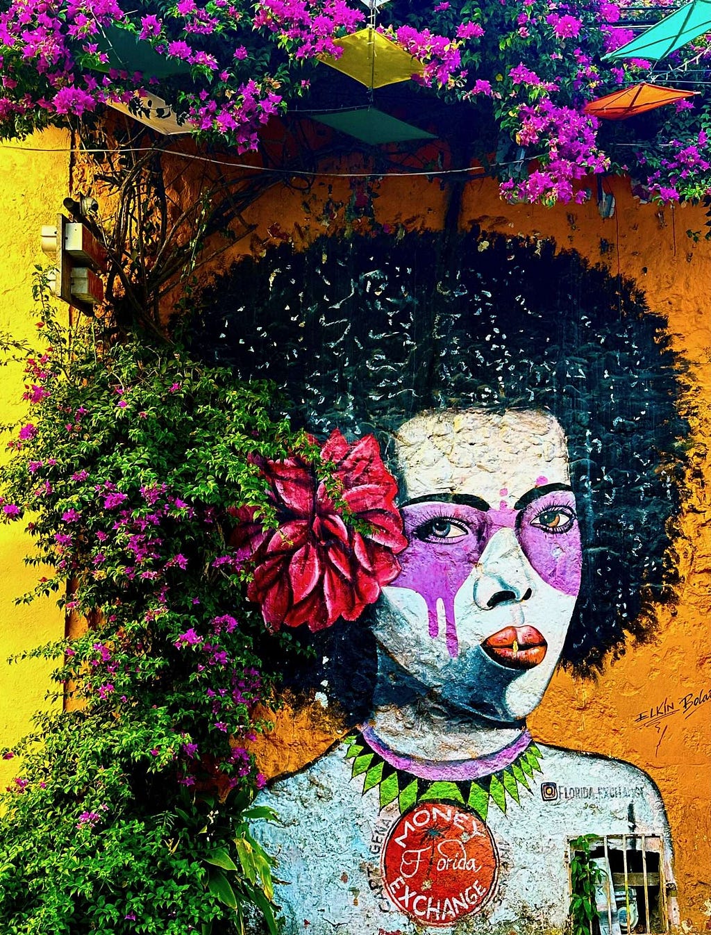 Colorful street mural in Getsamani depicting a woman with a flower in her hair and a purple face, surrounded by blooming bougainvillea on a yellow wall.
