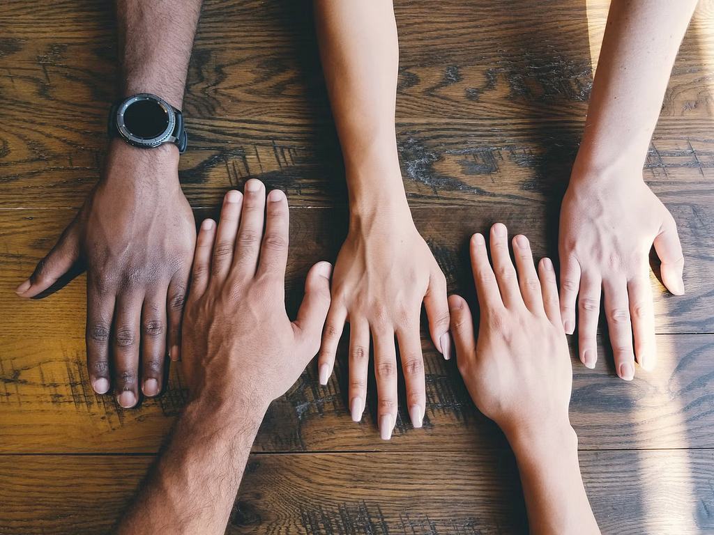 Photo of five hands (of five different people) with different skin complexions faced down side by side on a wooden table.