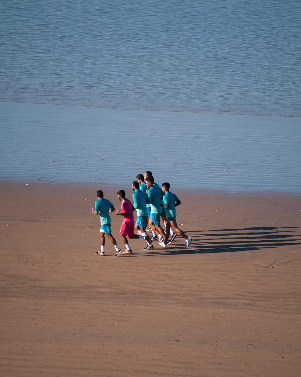 A group of men running on the beach. One man is dressed in red, the others in blue.