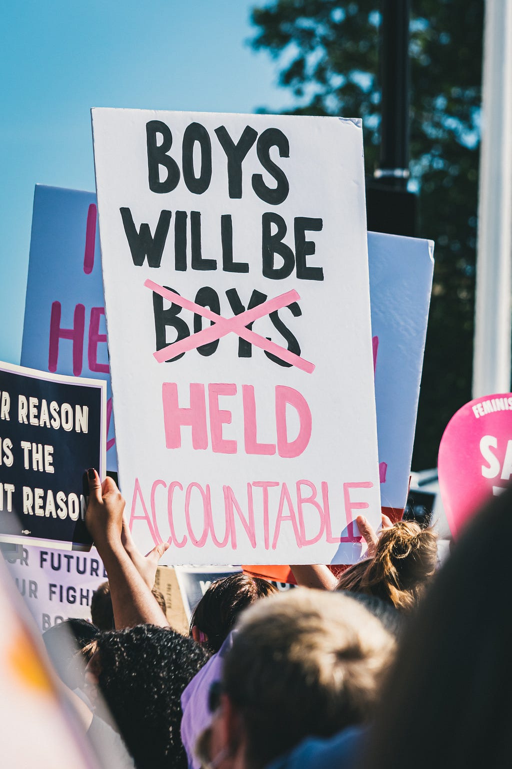 A protest sign in a forest of protest signs, emblazoned with “Boys will be held accountable” against a brilliant blue sky. Manicured hands thrust the sign skyward in defiance.