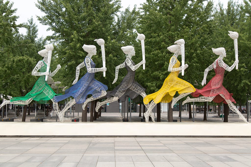 five wire sculptures of women dressed in green, blue, black, yellow, and red, each carrying the Olympic torch.