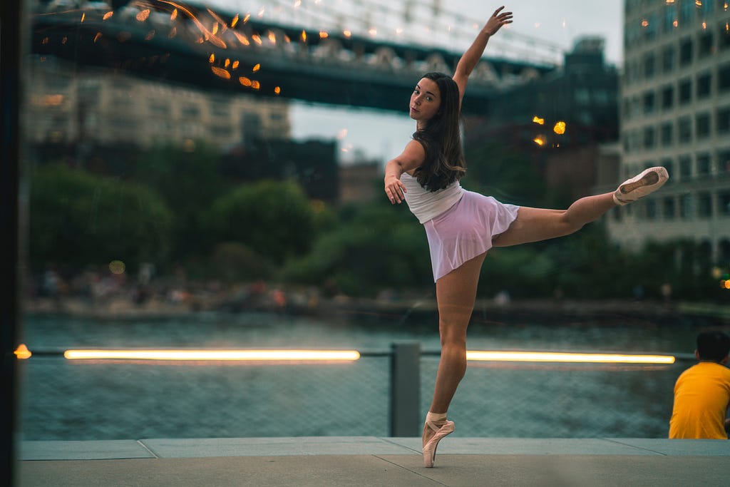 A young ballet dancer wearing white and pink in pointe shoes. She poses outside in an attitude pose and looks at the camera.
