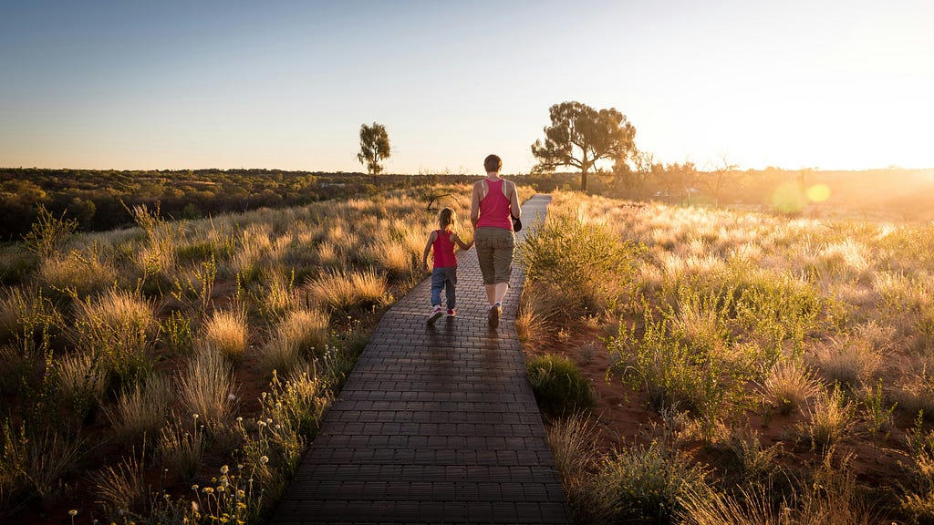 An image of a mother holding her son’s hand as they walk along a brightly lit path in the middle of a field.