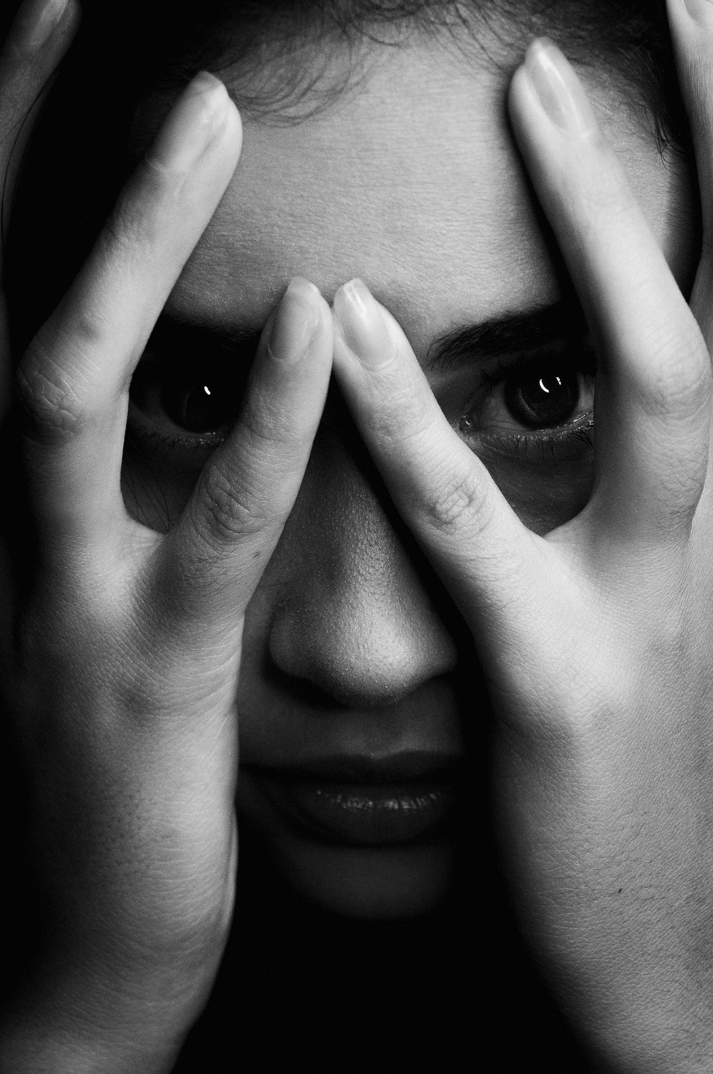 A young light skinned girl stares at the camera while covering her face with her hands, though her eyes are visible between the gaps in her fingers.