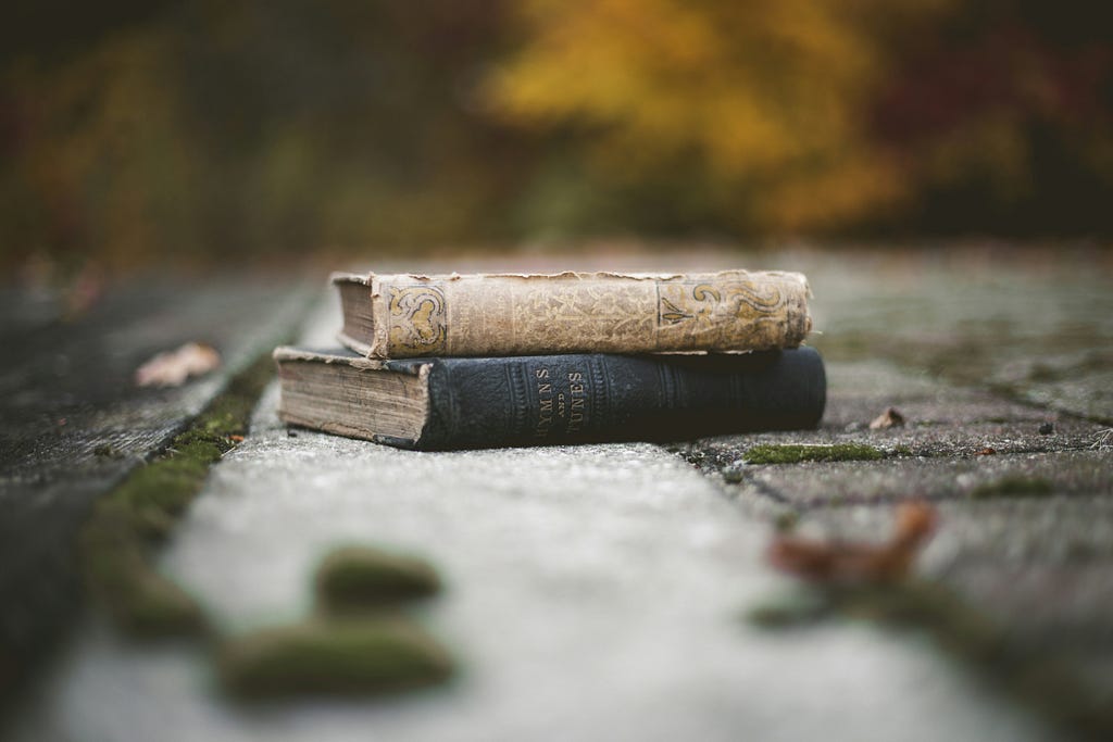 color photograph of two well-worn hardcover books laying outdoors on a walkway covered lightly in moss and crisp fallen leaves