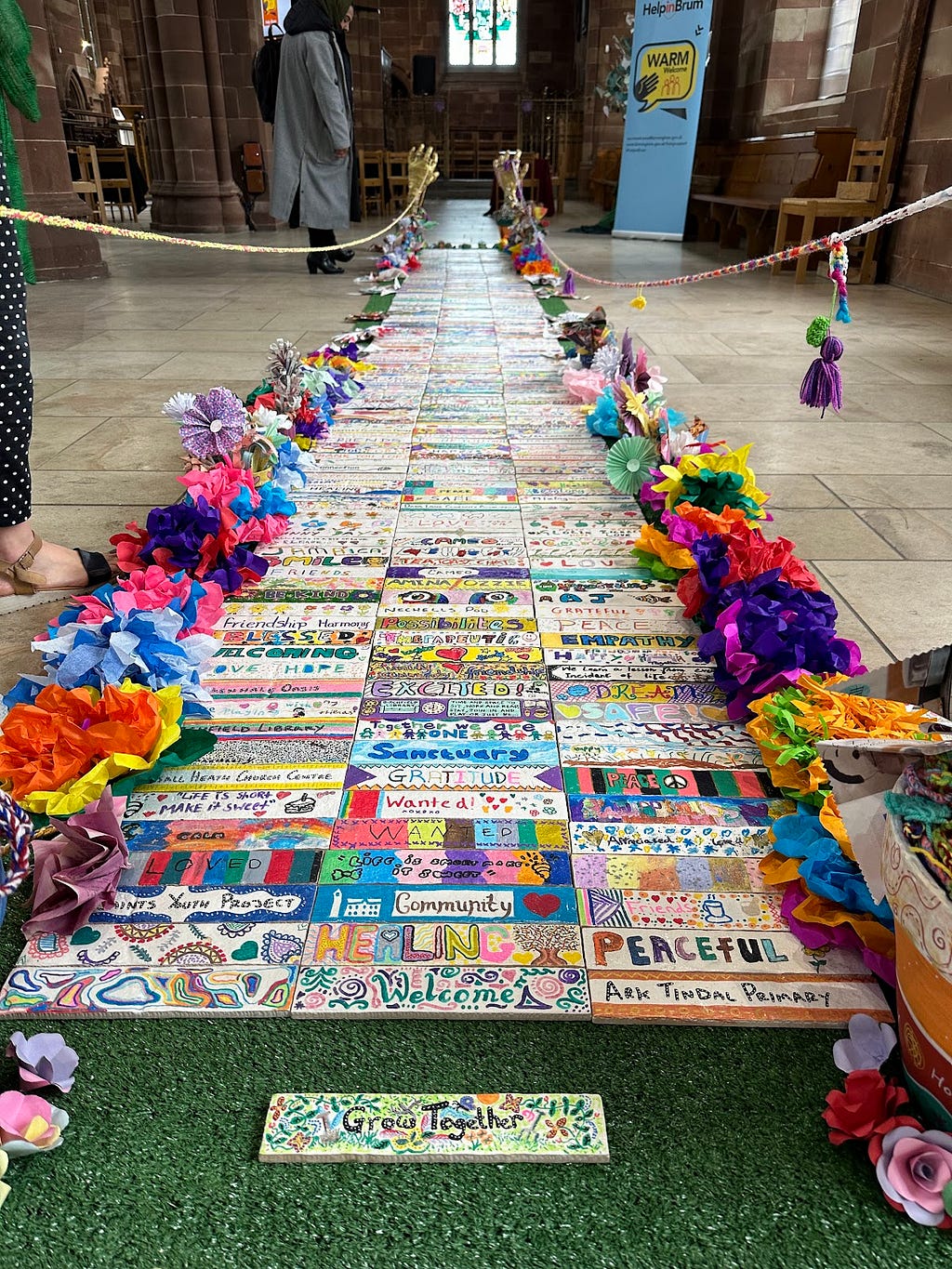 Warm Spaces guests in Birmingham wrote on tiles what the Spaces meant to them. This photo shows the tiles lined up on a church floor. It includes words like ‘wanted’, ‘community’, and ‘healing’. The tiles are colourful and flanked by flowers.