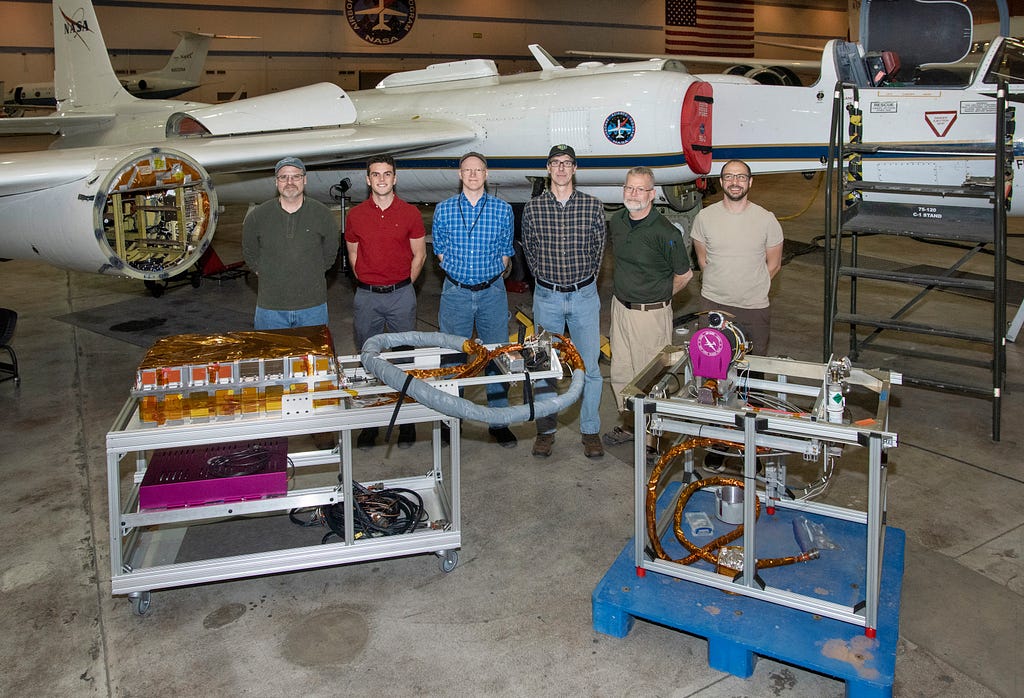 Six men pose in front of an airplane in a hangar with carts of equipment displayed in front of them.