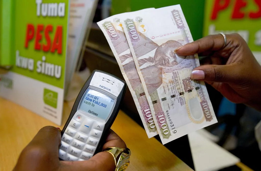 A mobile phone is shown beside physical East African currency in shillings behind the backdrop of M-pesa: a telecommunication company offering mobile money payments to consumers.